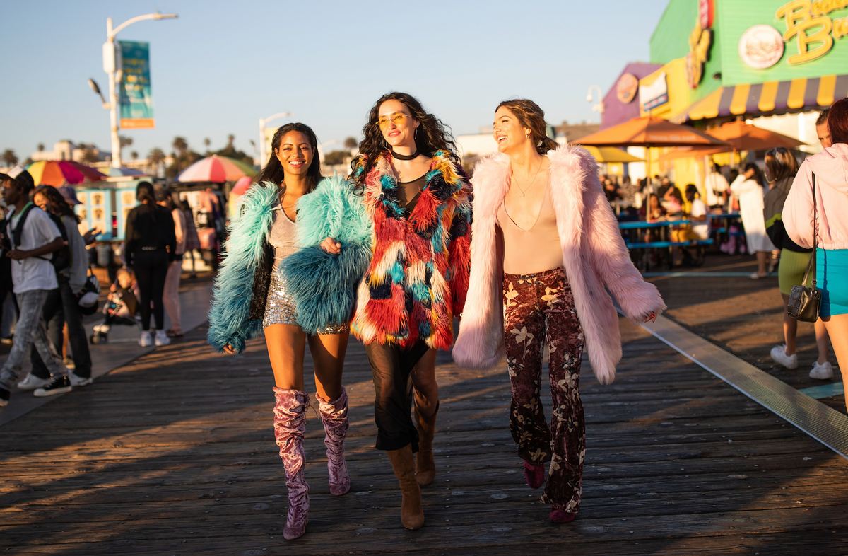 Three women dressed in colorful faux fur coats walk down a pier during a sunny day.