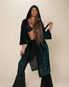 A woman wearing an emerald green velvet kimono with a hood, paired with black pants. The kimono has a sheerness and is designed with a tie belt at the waist and side seam pockets.