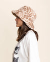 Person wearing a patterned tan leopard print fuzzy bucket hat and a white shirt with cat designs on a beige background