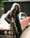 Male Model with short beard and mustache wearing hooded grey wolf vegan fur coat with hand in the pocket while leaning up again coral colored car outside.