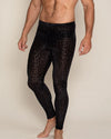 Man wearing black leopard print leggings on a beige background - leggings are a sheer black with burnout velvet black leopard print spots throughout