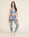 A woman models metallic silver leggings with a multicolor holographic foil snakeskin pattern. She is also wearing a matching top and white sneakers.