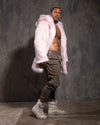 A man wearing a pink fake fur coat, gray pants, and white boots against a textured gray wall.