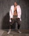 Man wearing a pink fake fur coat, gray pants, and white sneakers against a dark textured wall.