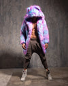 Man wearing wearing a colorful tie dye pink and blue faux fur coat with oversized hood and bear ears, against a textured wall.