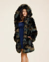 Woman wearing a thigh length, midlength fake fur coat in green and brown camo inspired fake fur coat with a hood on a beige background