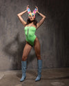 A Woman wearing a green swimsuit, and a pastel tie dye mask, with bunny ears, and blue boots against a textured gray wall.