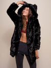 Female model gazing upward and smiling  while holding ear on hood of the fake fur black panther jacket.