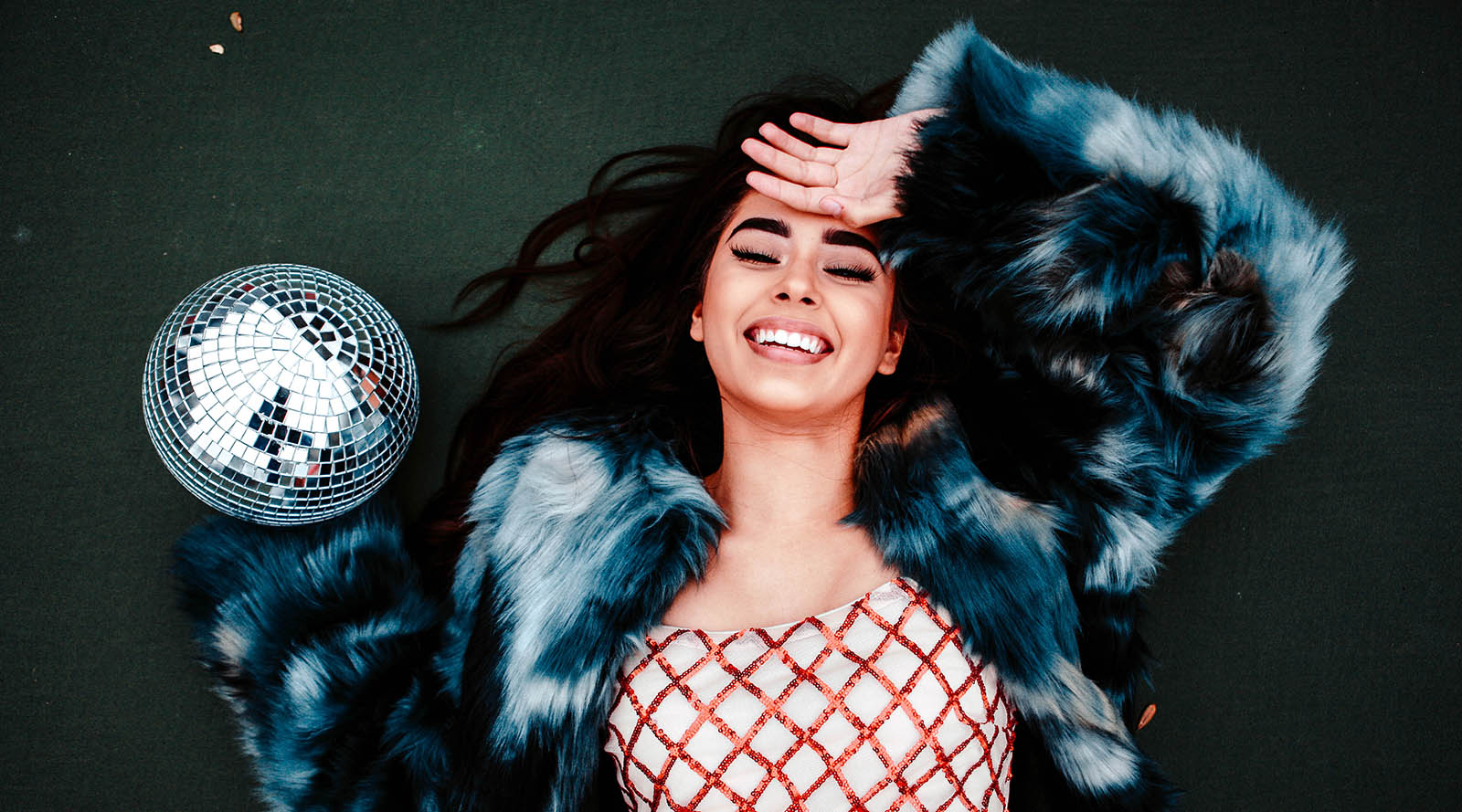 girl lying down with hand over forehead wearing faux fur coat, red and white shirt, and disco ball