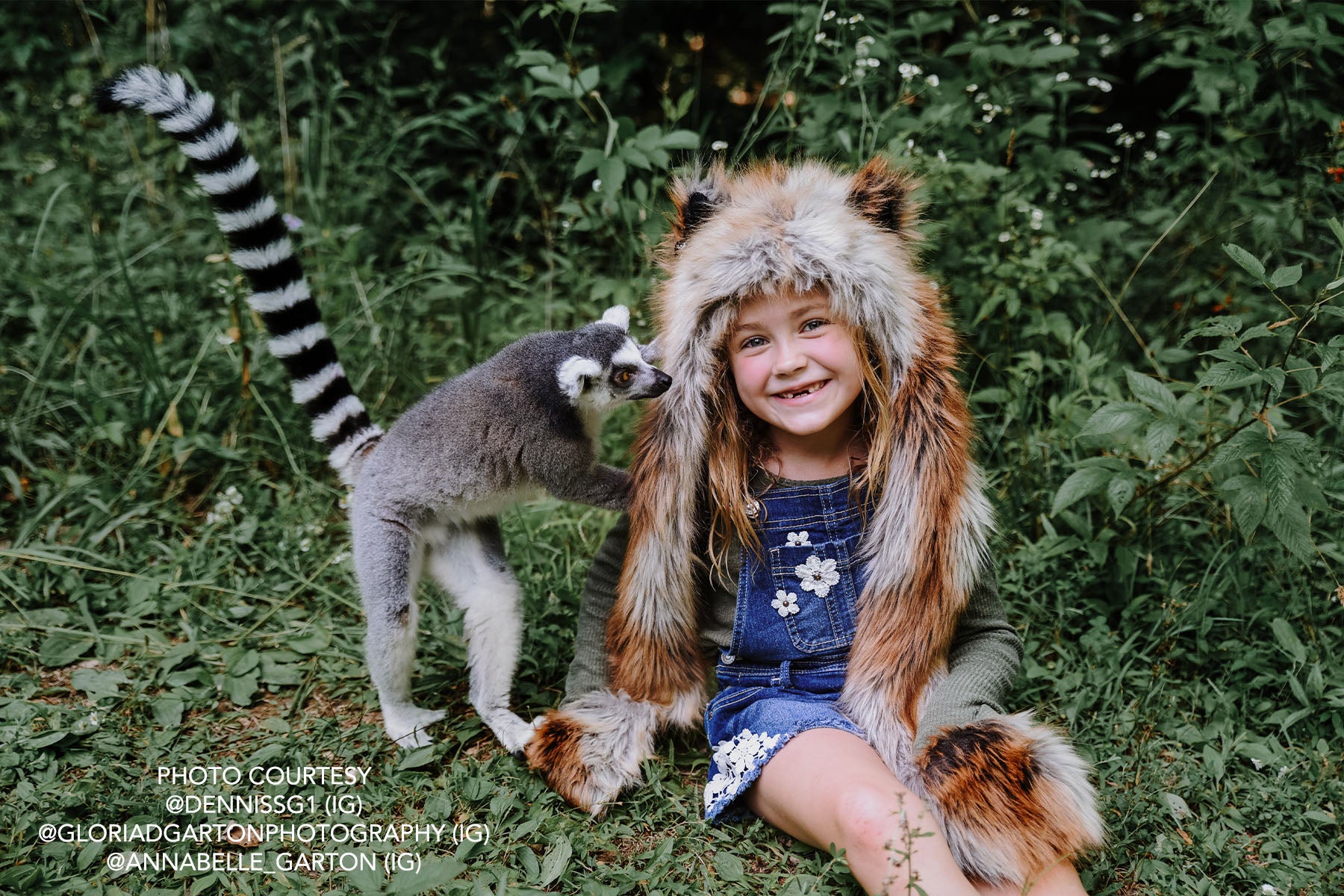 girl wearing faux fur hood sitting in grass with lemur