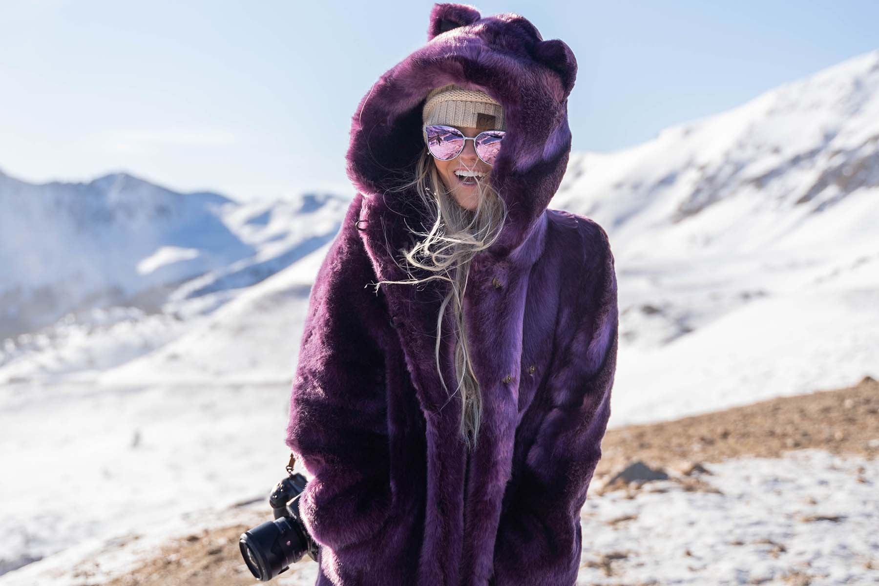 woman standing in snowy mountains wearing purple faux fur coat