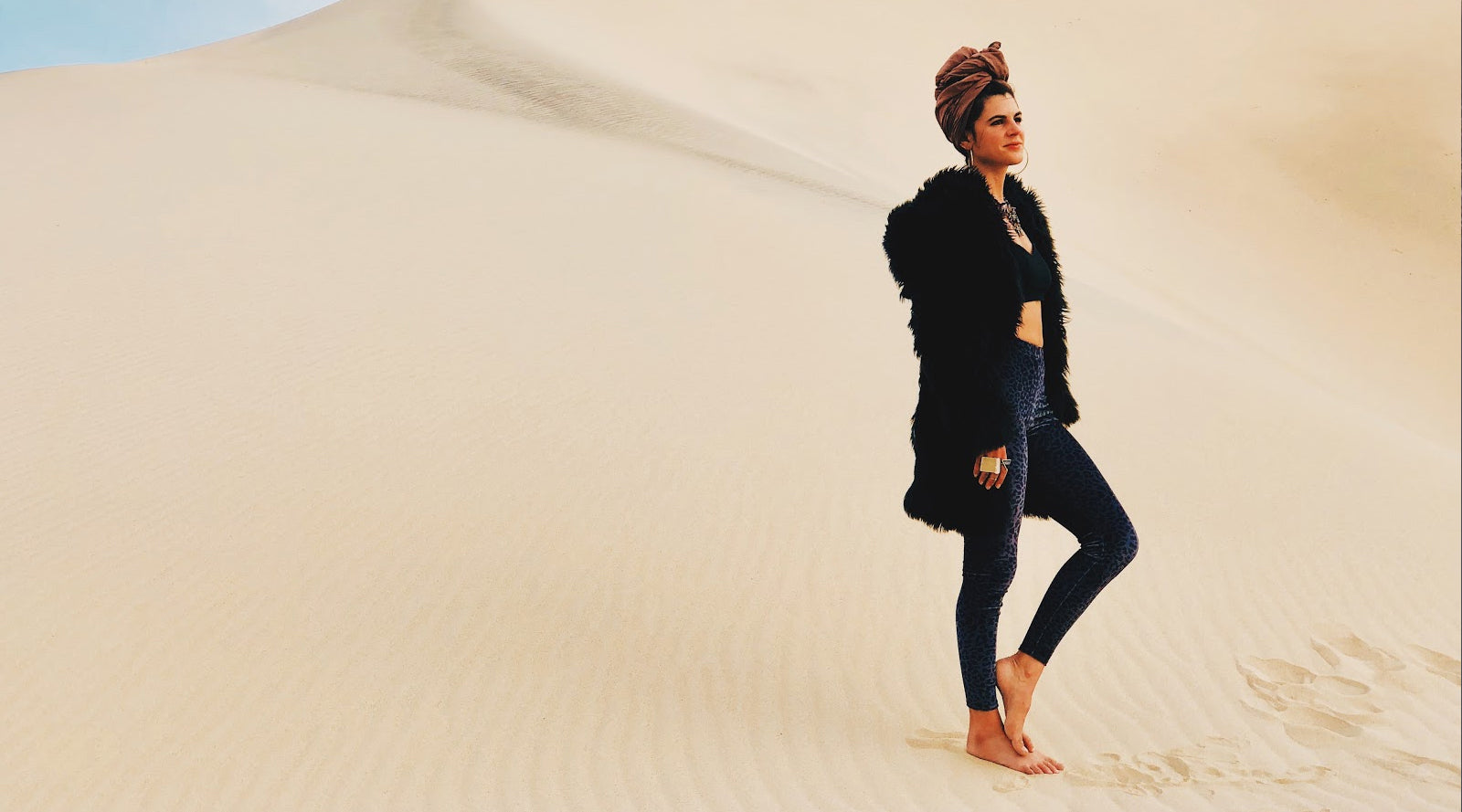 person wearing headband, leggings, and faux fur barefoot in sand