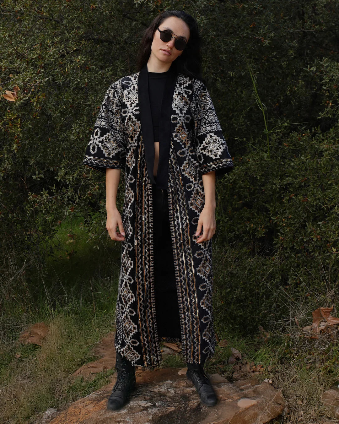 A model wearing a long black kimono-style cardigan with intricate white and gold geometric patterns, styled over a black outfit and standing outdoors on a rock in front of a dense green forest.