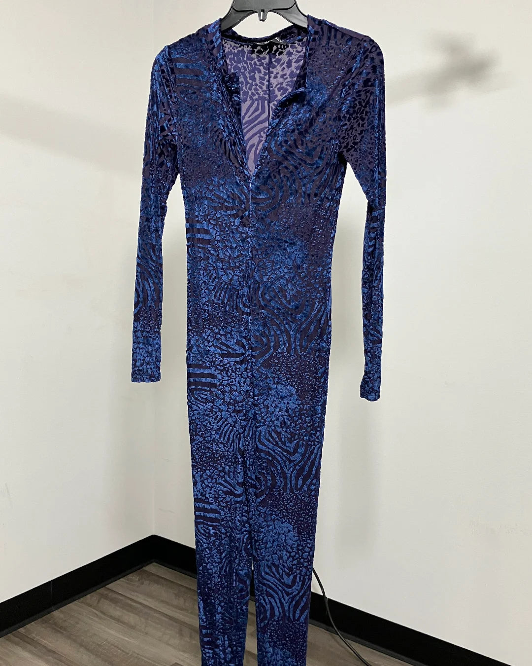 A long-sleeve, full-length jumpsuit with a blue and purple abstract pattern, hanging on a black hanger against a plain off-white wall.