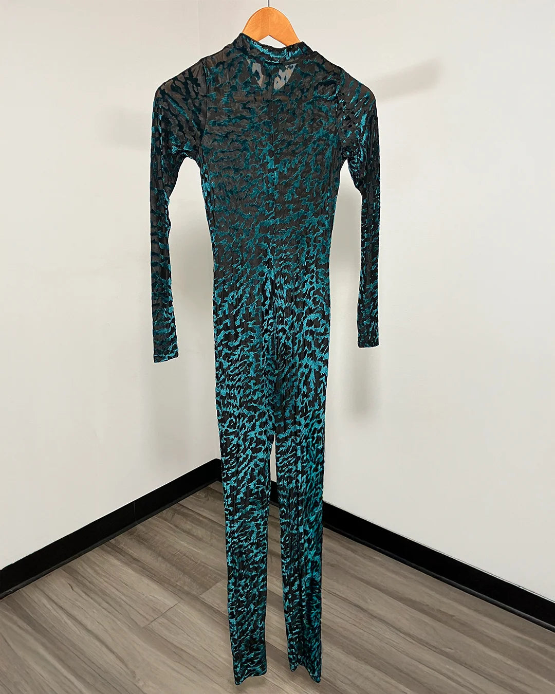 A long-sleeve full-length jumpsuit with a black and teal-green leopard print pattern, hanging on a wooden hanger against a white wall.
