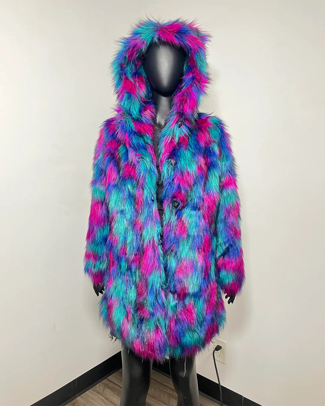 A hooded faux fur coat in a vibrant pink, blue, and teal pattern, displayed on a black mannequin against a plain off-white wall.