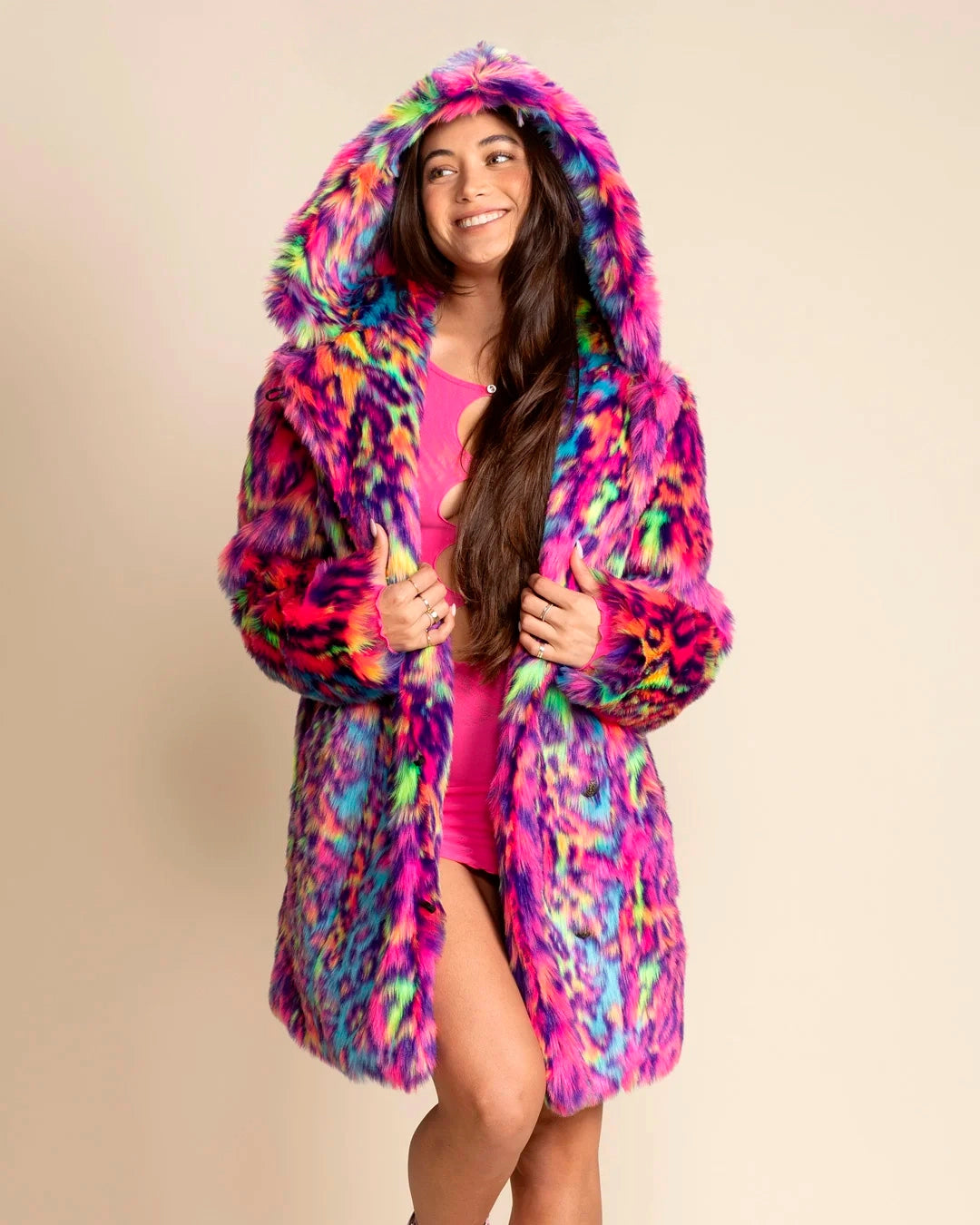 A Woman wearing a colorful neon pink, blue, green leopard print fake fur coat, with a large oversized hood, as she smiles facing away from the camera against a beige background. SpiritHoods Fall Winter Custom Made Faux Fur Coats and Animal Print Jackets that Glow under UV Blacklight