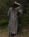 A woman viewed from behind wearing a long bohemian-style patterned robe with wide sleeves, standing outdoors in front of a dense green forest. The robe features intricate geometric and tribal patterns in black, white, and brown.