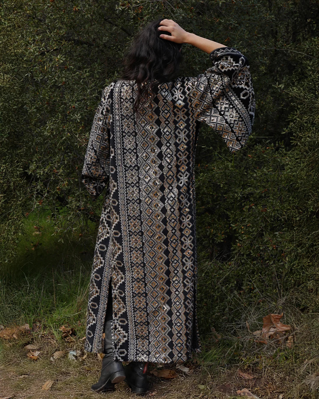A woman viewed from behind wearing a long bohemian-style patterned robe with wide sleeves, standing outdoors in front of a dense green forest. The robe features intricate geometric and tribal patterns in black, white, and brown.