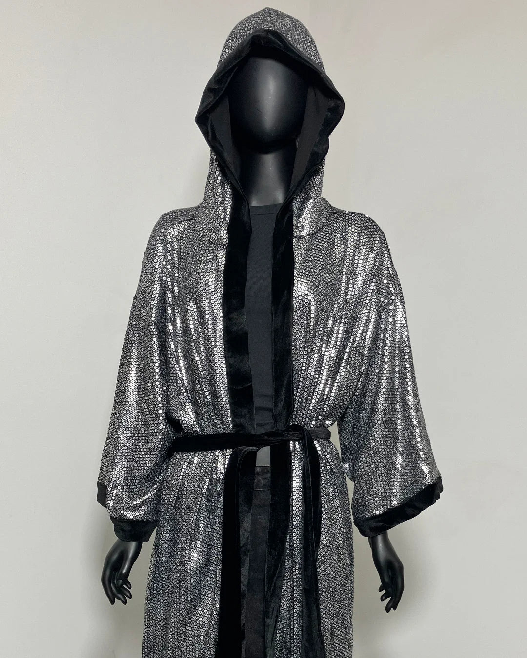 A hooded robe with a silver sequined exterior and black satin trim, displayed on a black mannequin against a plain light gray background.