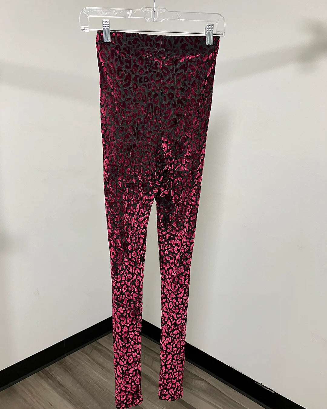 Magenta pink and black leopard print leggings hanging on a clear plastic hanger against a plain white wall.