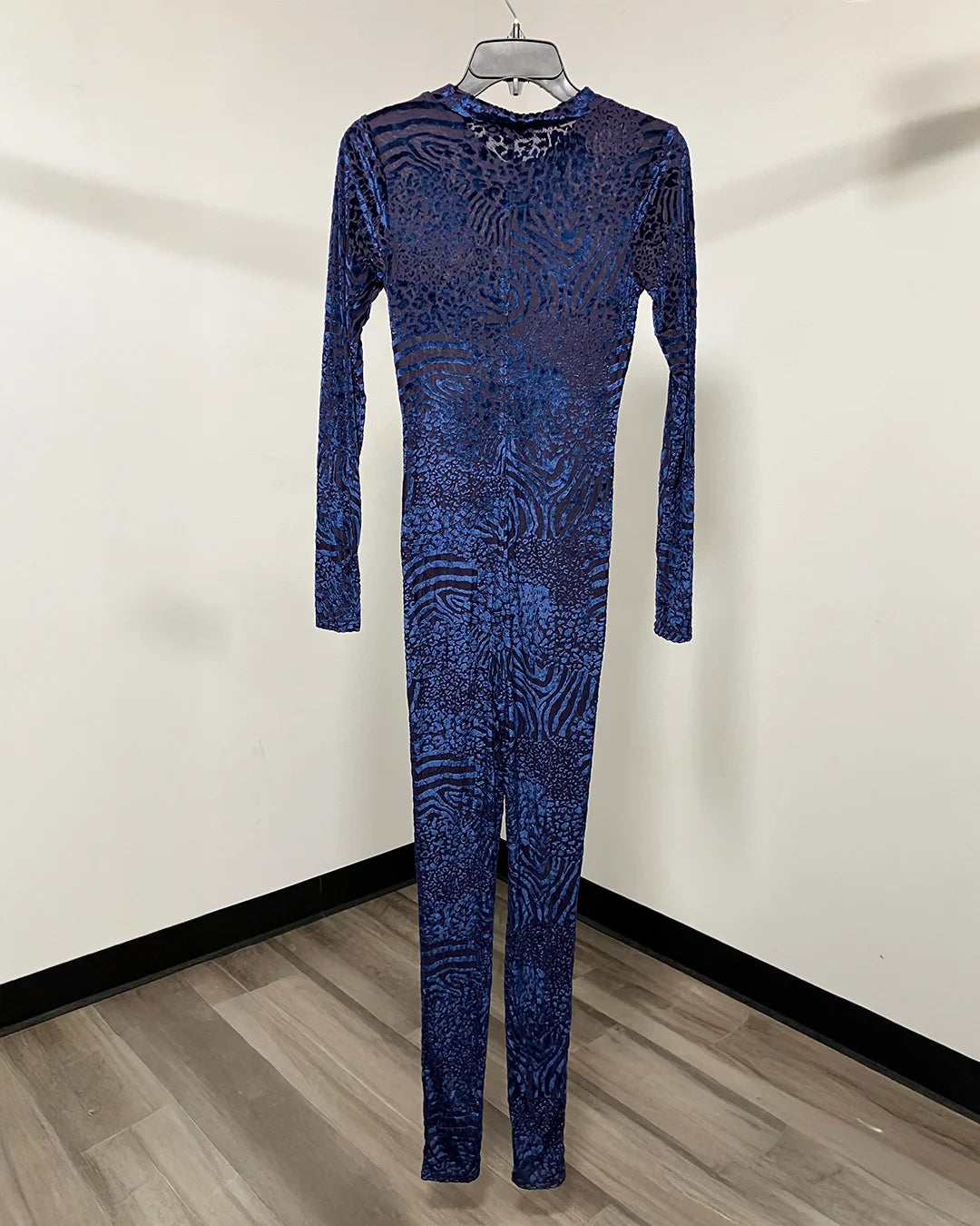A long-sleeve full-length one-piece jumpsuit with an abstract blue and black pattern, hanging on a hanger against a plain off-white wall with a wood-look floor.