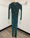 A full-body one-piece jumpsuit with a long sleeve and front zipper, hanging on a wooden hanger against a plain white wall. The fabric has an abstract black and teal-green pattern.