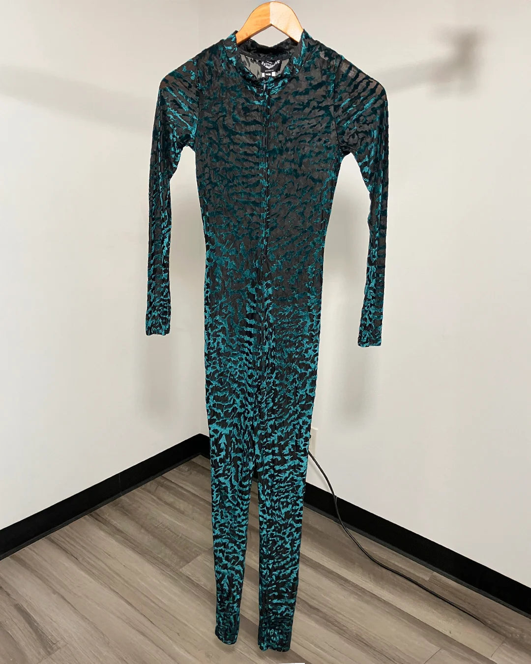 A full-body one-piece jumpsuit with a long sleeve and front zipper, hanging on a wooden hanger against a plain white wall. The fabric has an abstract black and teal-green pattern.