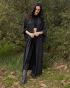 A woman stands outdoors in a wooded setting wearing a long black textured crochet cardigan over a black outfit, paired with black knee-high boots.