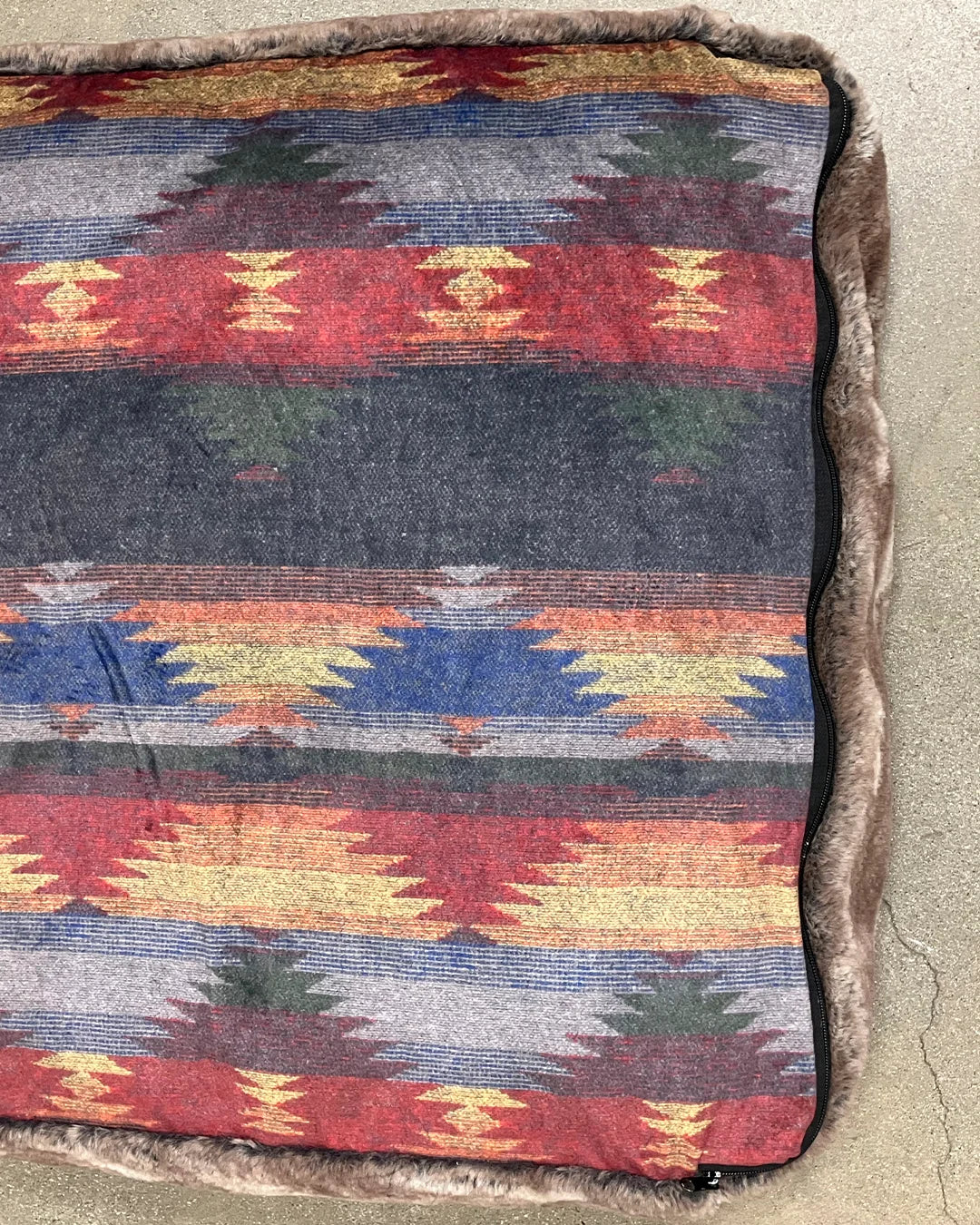 Multicolored patterned pet bed with a textured border on a concrete floor.