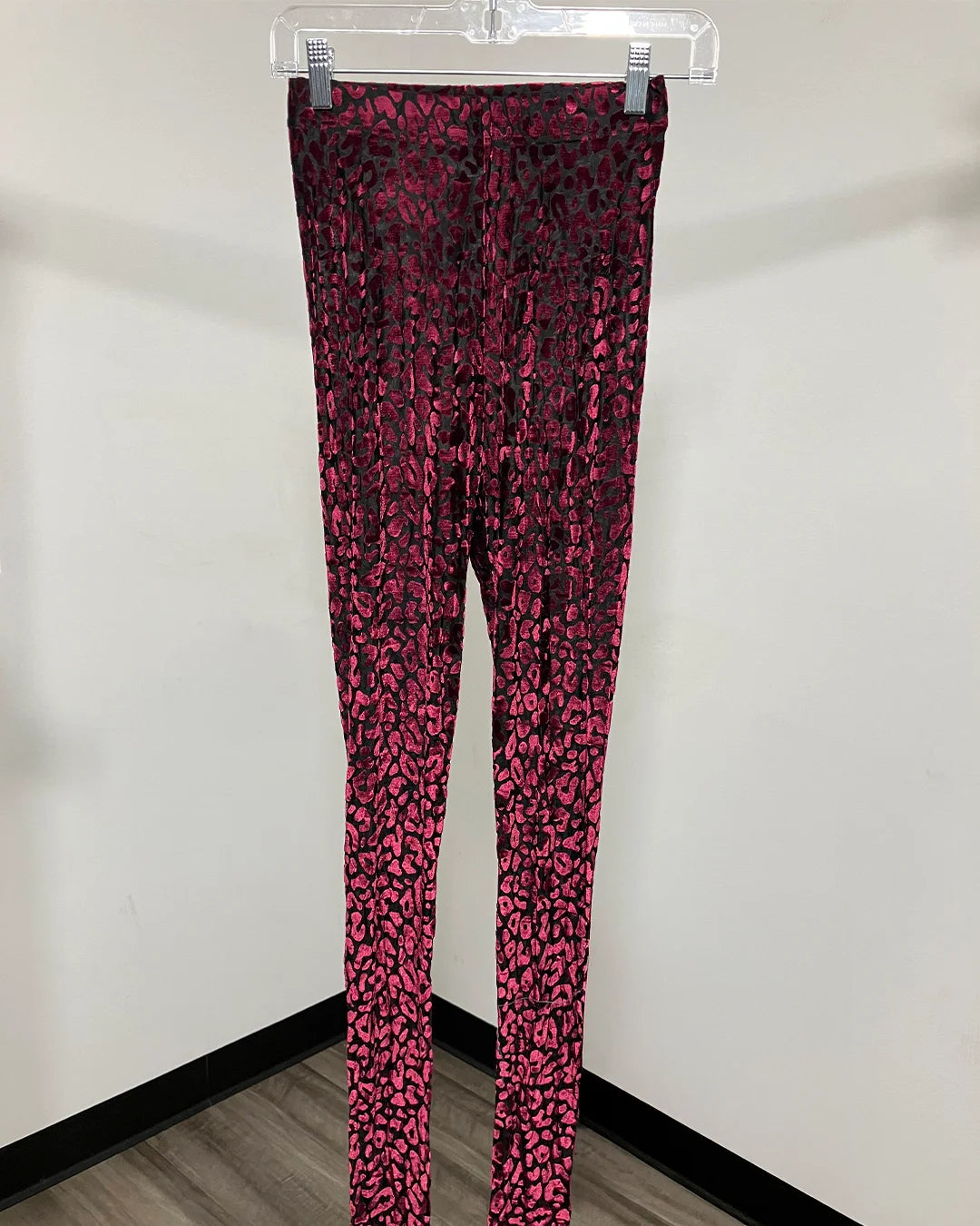 Red and black leopard print leggings hanging on a clear plastic hanger against a plain white wall.