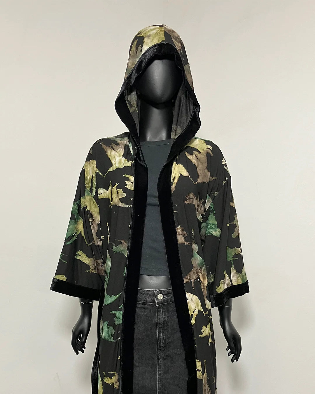 A hooded longline kimono-style jacket with a green and brown hummingbird silhouette pattern and black trim, displayed on a black mannequin against a plain light background.