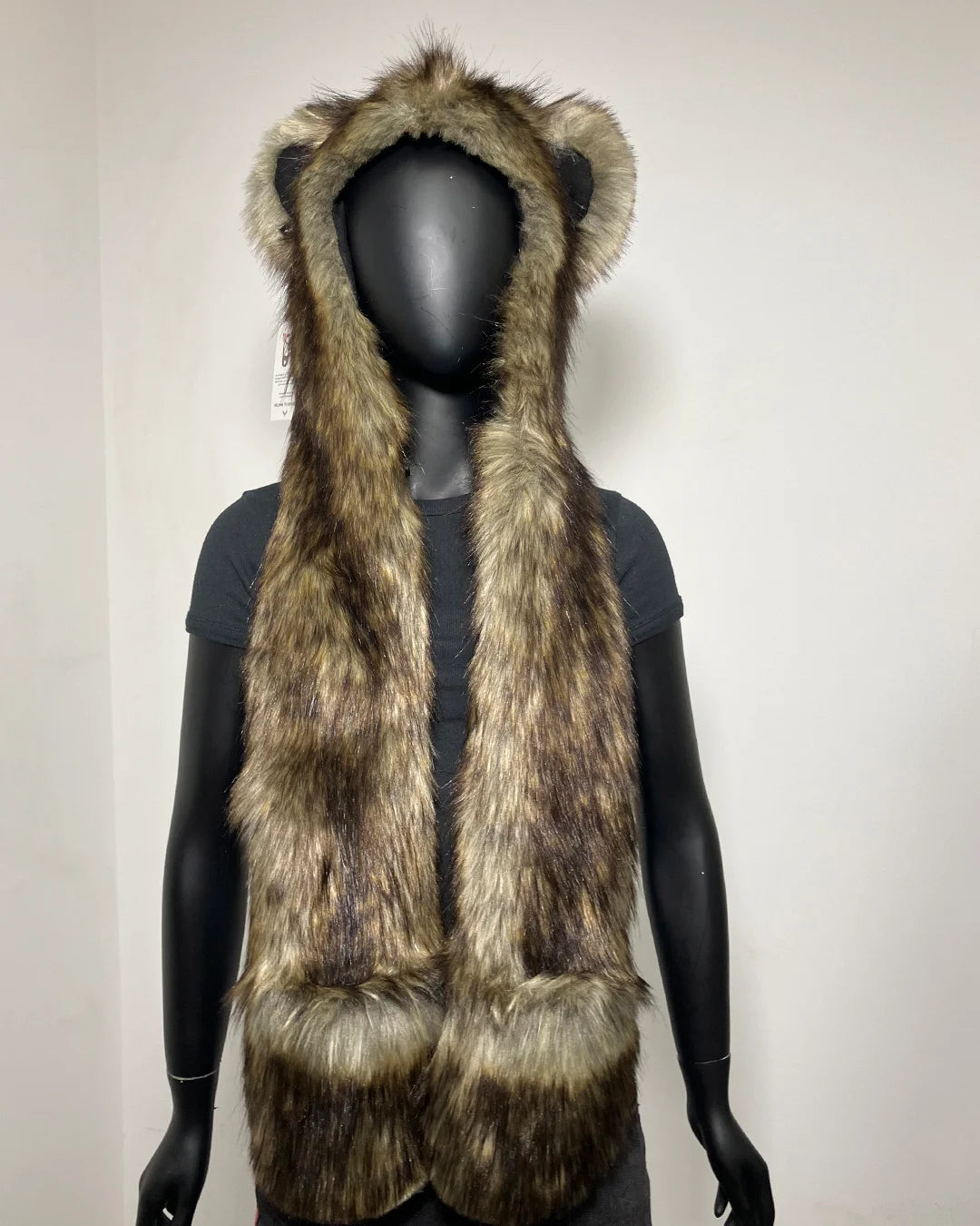 A brown and beige faux fur hood with animal ears, displayed on a black mannequin torso against a plain light gray wall.
