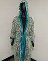 A hooded robe covered in iridescent sequins with teal velvet trim and a matching tie belt, displayed on a black mannequin against a plain indoor wall.