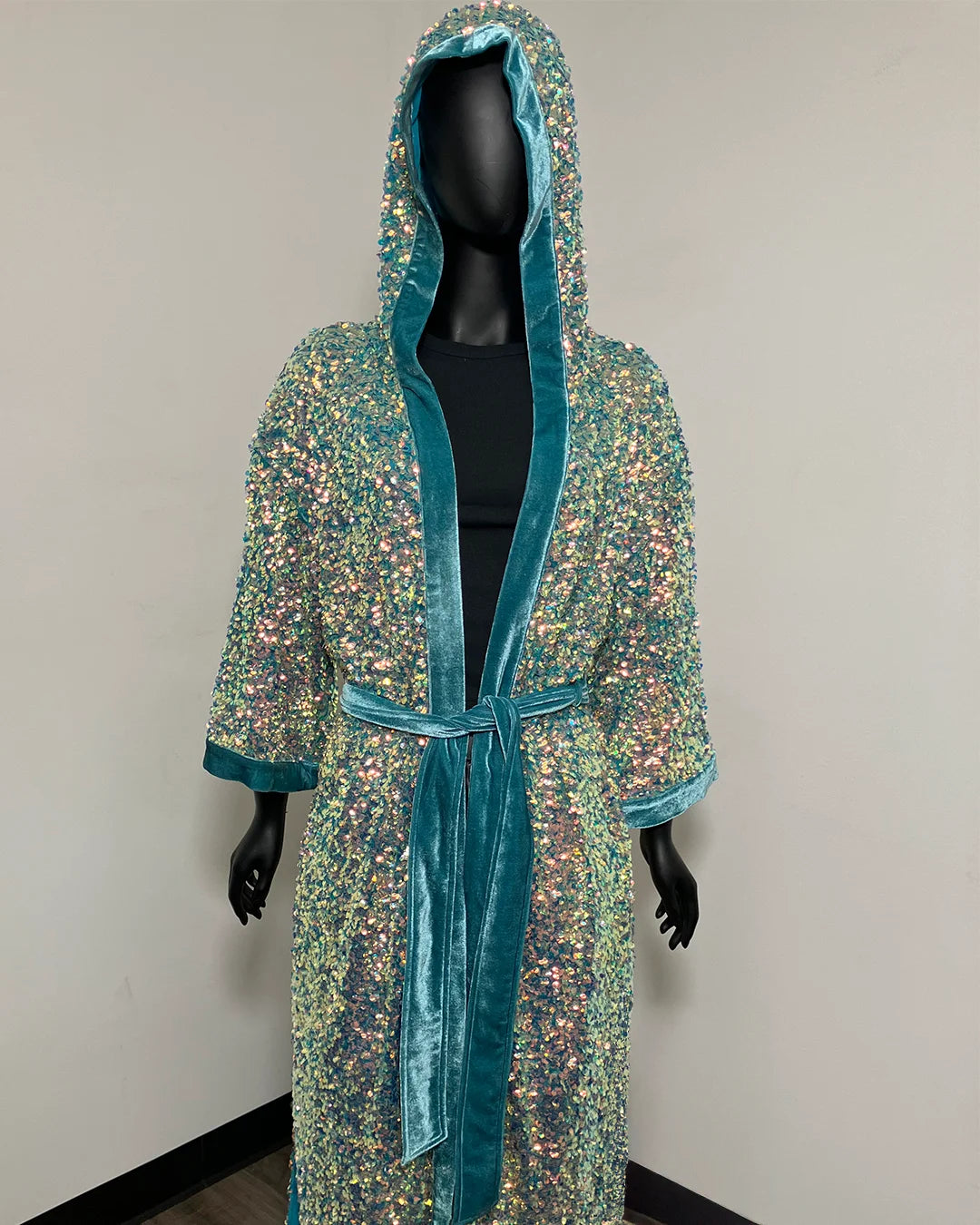 A hooded robe covered in iridescent sequins with teal velvet trim and a matching tie belt, displayed on a black mannequin against a plain indoor wall.