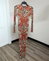 A long-sleeve bodysuit with a bold abstract pattern in red, beige, and black, hanging on a black hanger against a white wall and wood-look floor.