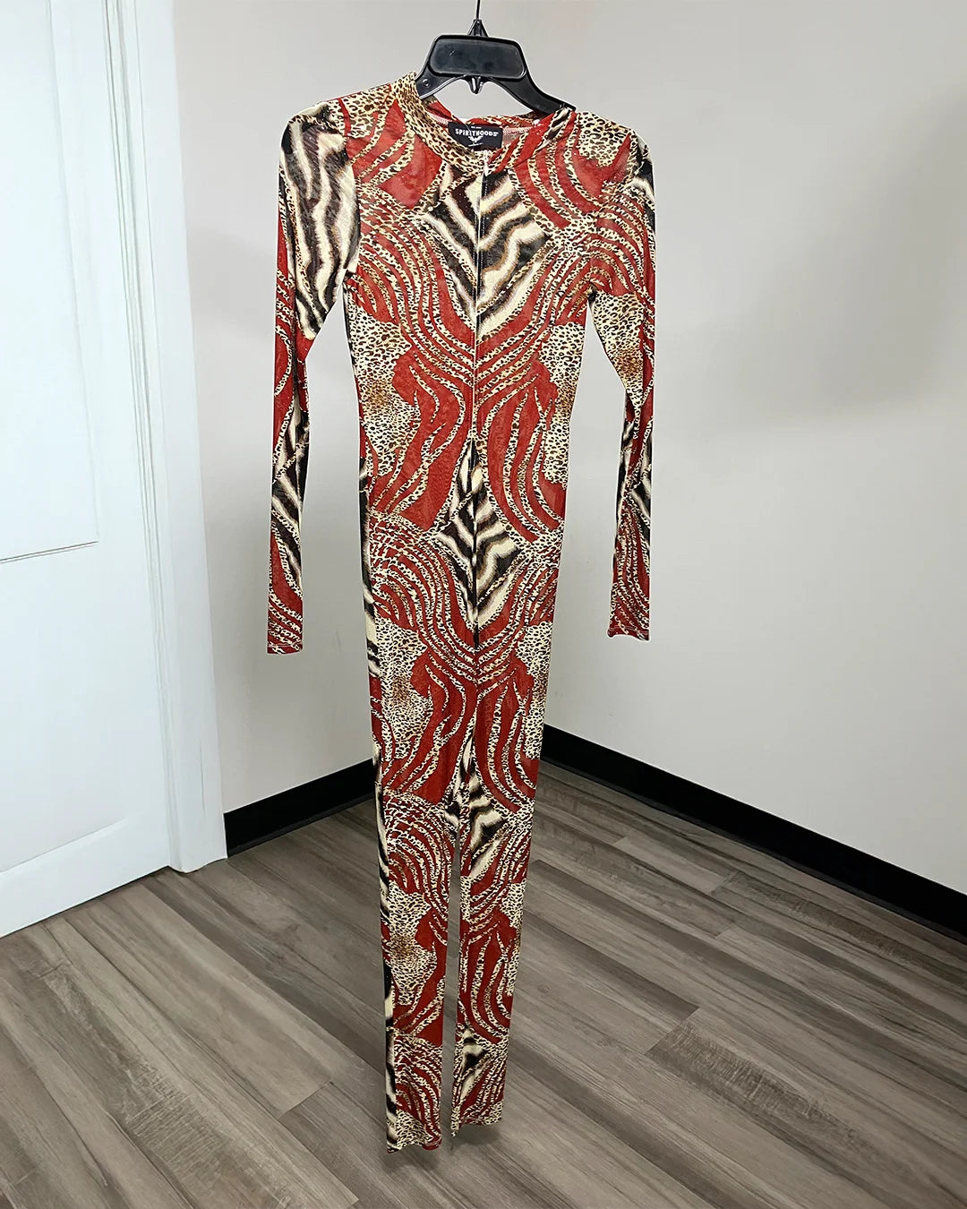 A long-sleeve bodysuit with a bold abstract pattern in red, beige, and black, hanging on a black hanger against a white wall and wood-look floor.