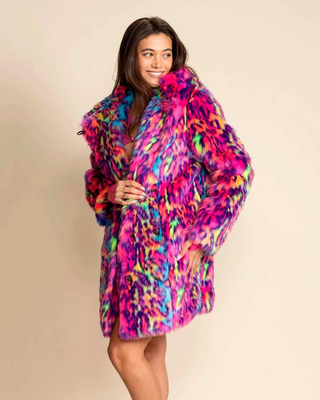 A Woman wearing a colorful neon pink, blue, green leopard print fake fur coat, with a large oversized hood, as she smiles facing away from the camera against a beige background. SpiritHoods Fall Winter Custom Made Faux Fur Coats and Animal Print Jackets that Glow under UV Blacklight