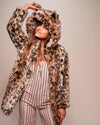 A Woman wearing a leopard print coat with a hood, pulling up on the ear and smiling, against a plain background. SpiritHoods Fall Faux Fur Coats and Custom Made Fake Fur Jackets for Women