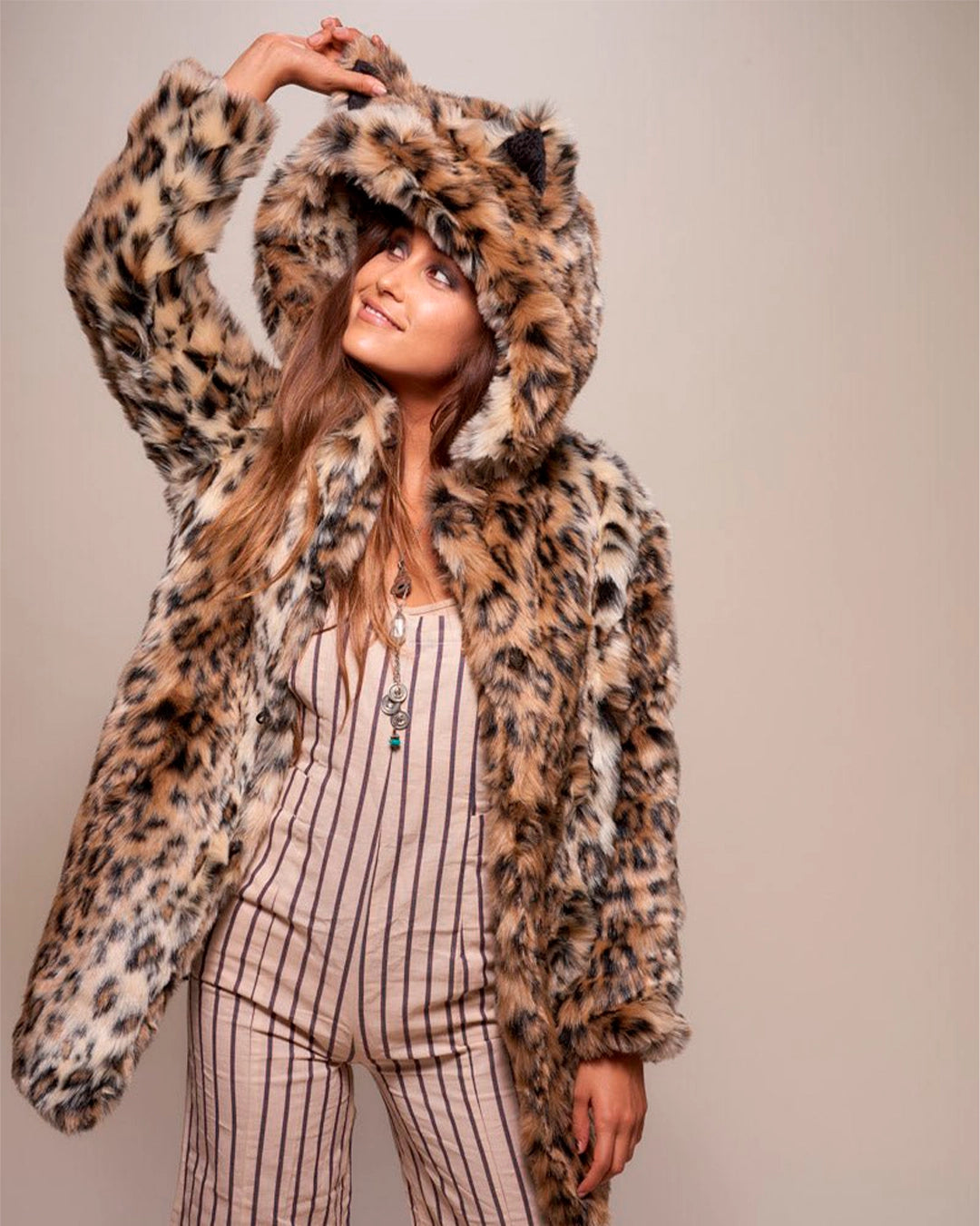 A Woman wearing a leopard print coat with a hood, pulling up on the ear and smiling, against a plain background. SpiritHoods Fall Faux Fur Coats and Custom Made Fake Fur Jackets for Women