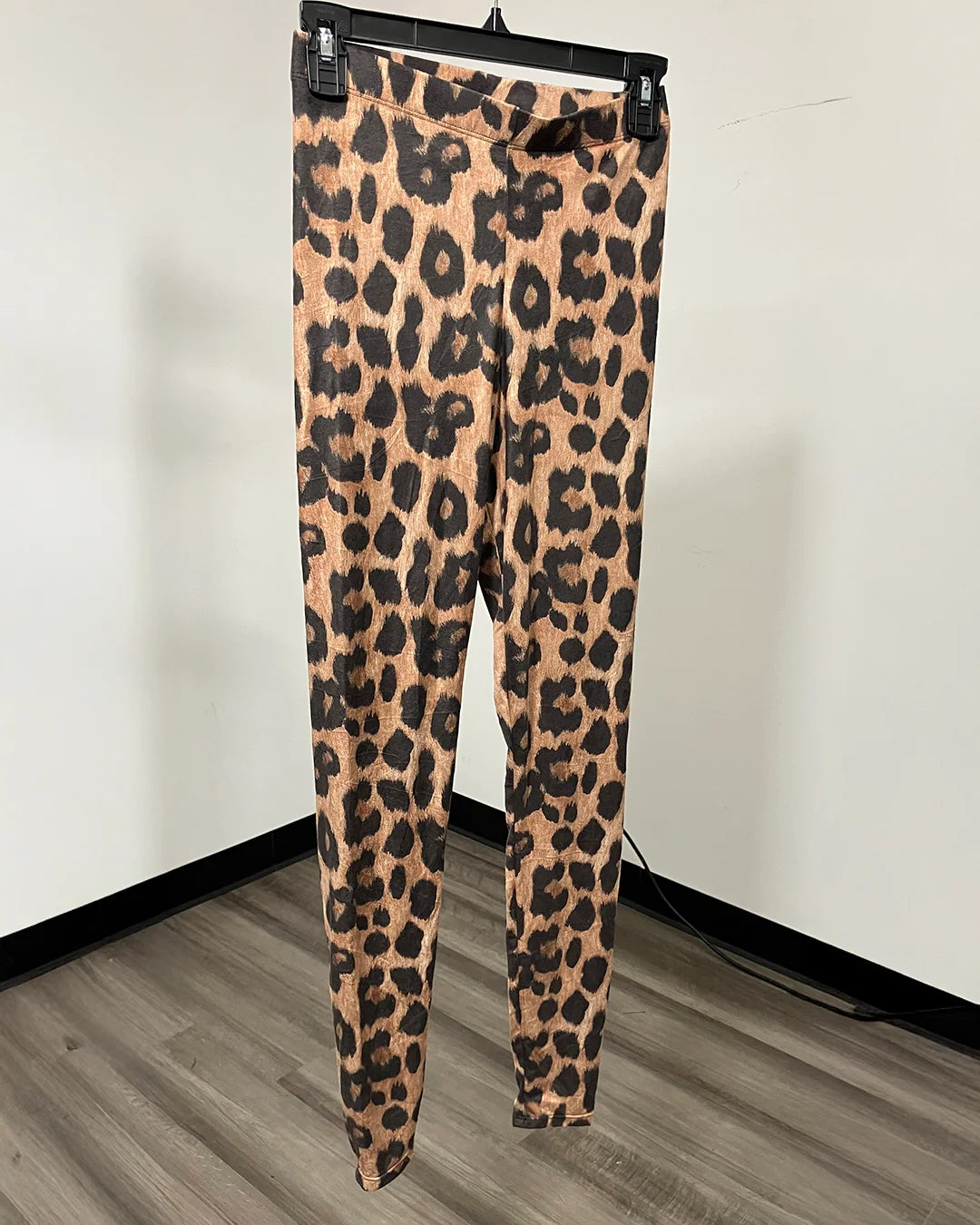Leopard print leggings hanging on a black hanger against a white wall and wood floor.