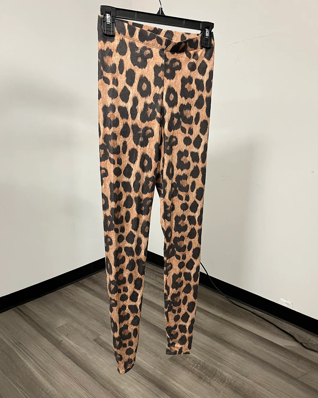 Leopard print leggings hanging on a black hanger against a white wall and wood-look floor.