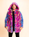 A Man wearing a colorful fur-free fur coat against a beige background. Neon Pinks, Neon Blue, Neon Yellow leopard print throughout, that glows under UV Blacklight. Featuring SpiritHoods oversized Hood and leopard face pea-coat buttons.  Custom Made Coats and Made to Order Faux Fur Jackets