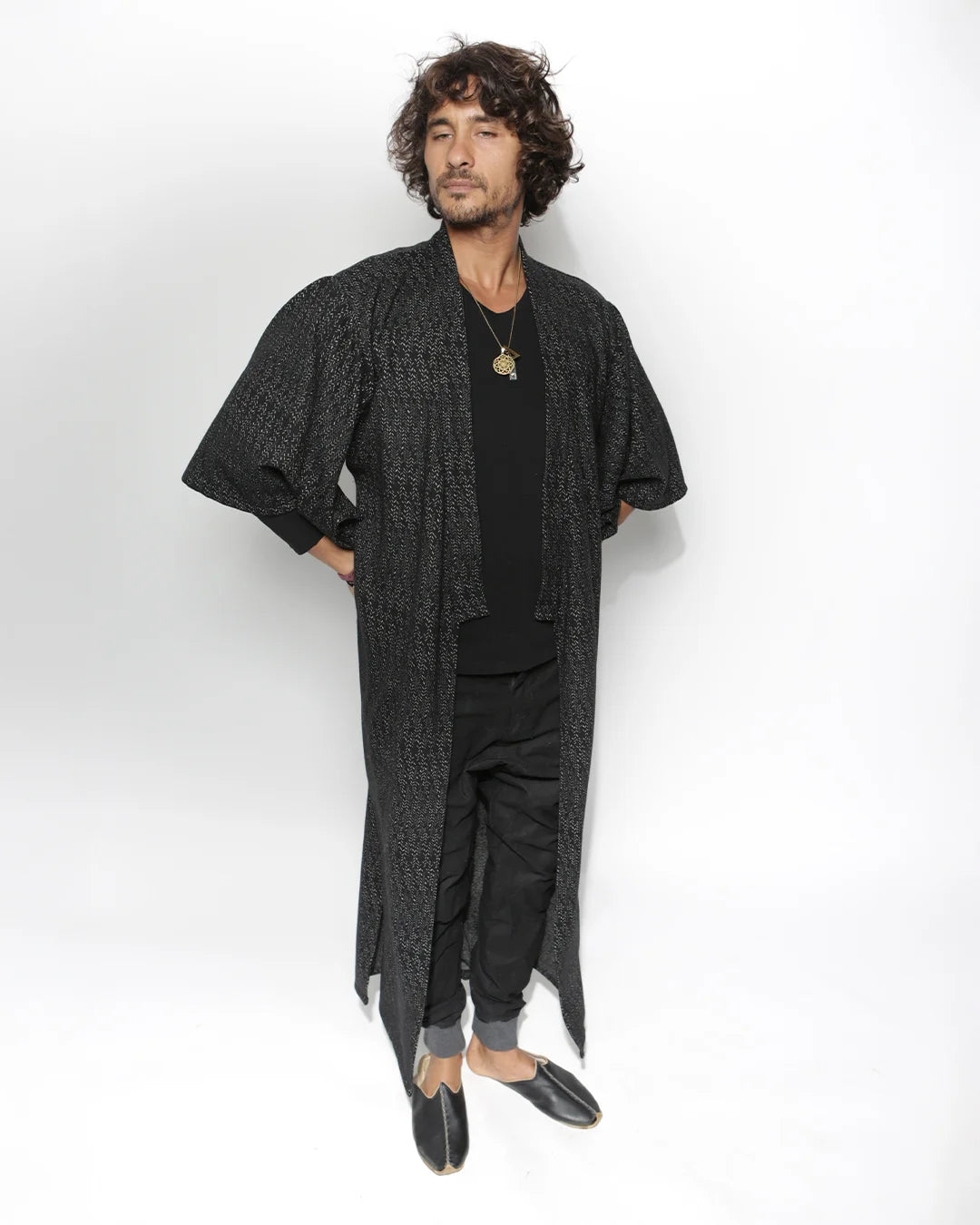 A man modeling a long, open-front black textured kimono-style robe over a black outfit, standing against a plain white studio background.