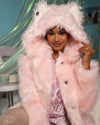 A Woman wearing a Pink Faux Fur Coat with an oversized hood and wolf ears on top, the coat is a light pink, and down the wearer's left side you can see a row of pea-coat style Wolf Face buttons. She is looking into the camera smiling, holding the left side of the hood. SpiritHoods 100% Fake Fur Coats and Womens Faux Fur Jackets