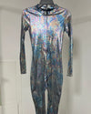 A long-sleeve, one-piece full-body suit with a high collar, displayed on a hanger against a plain light gray wall. The fabric has a strong iridescent, holographic finish reflecting blue, purple, and silver tones.