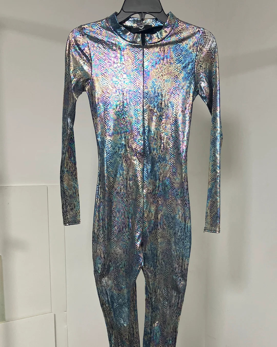 A long-sleeve, one-piece full-body suit with a high collar, displayed on a hanger against a plain light gray wall. The fabric has a strong iridescent, holographic finish reflecting blue, purple, and silver tones.