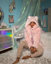 Person wearing a pink faux fur SpiritHoods coat and sitting on a bed in a room with a colorful dresser and decorative items.