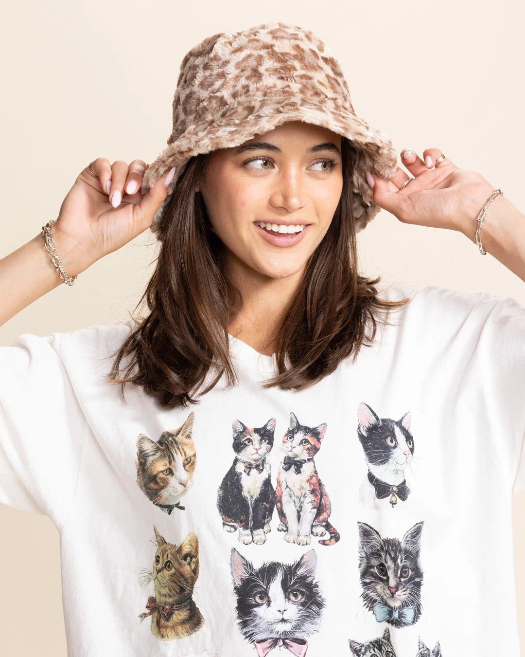 Woman wearing a tan leopard bucket hat and a white t-shirt with cat prints on a beige background