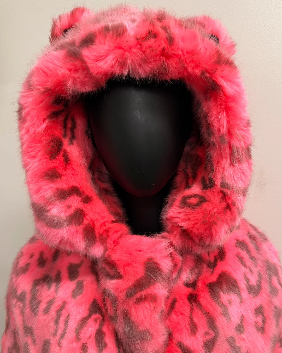 Mannequin wearing a bright pink faux fur coat with black leopard print pattern on a plain background. Showing the Smaller than standard ears on the top of the coats hood
